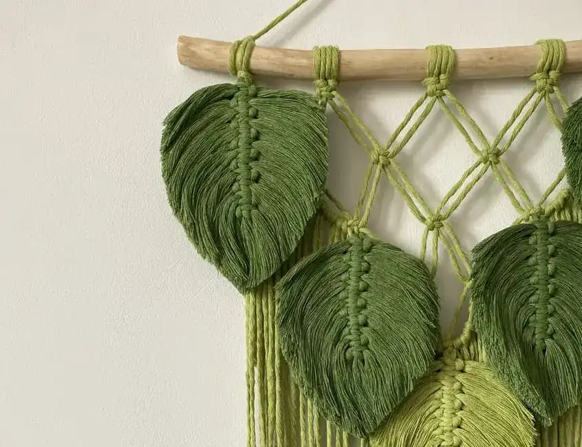 Green knotted leaves hanging from a wooden branch beside macramé tools